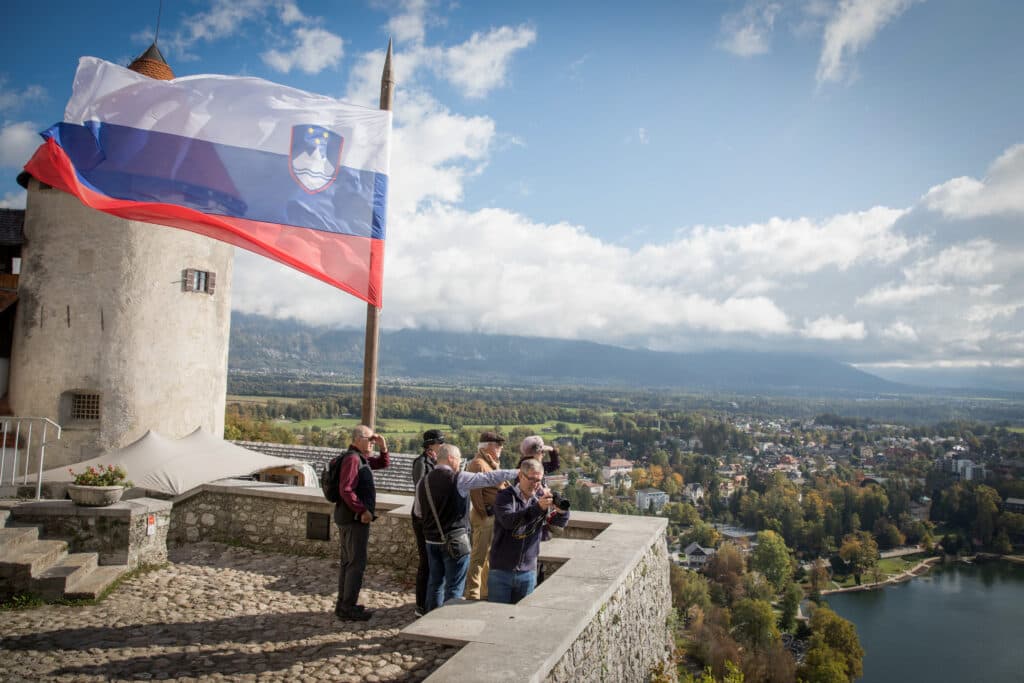 Tourists stand near a Slovenian flag at a lookout point over Lake Bled.