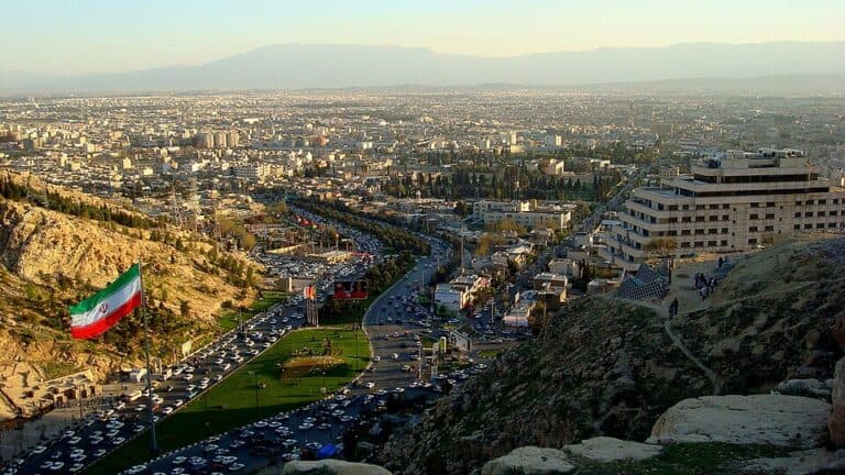 A wide view of Shiraz, Iran, with mountains, city streets, buildings, and an Iranian flag visible.