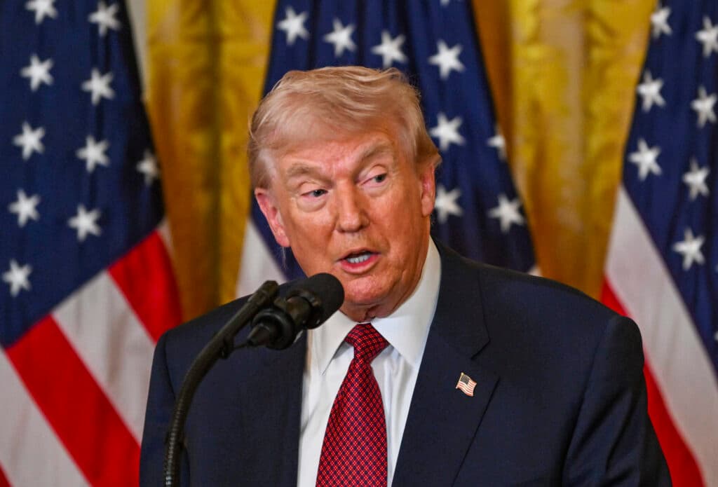 President Donald Trump speaks at a microphone in the East Room of the White House with American flags behind him.