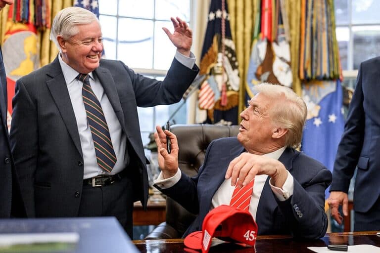 Donald Trump seated at the Resolute Desk with Lindsey Graham standing beside him in the Oval Office.