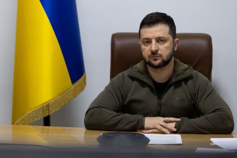 Ukrainian President Volodymyr Zelenskyy sits at a desk beside a Ukrainian flag.