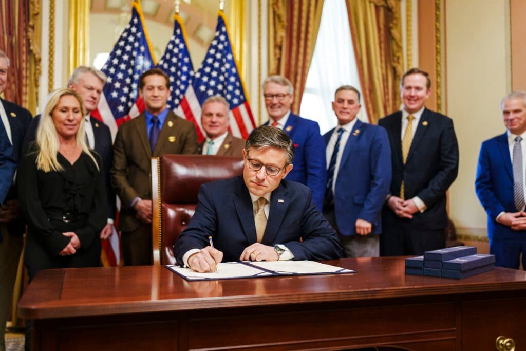 U.S. House Speaker Mike Johnson signs a document while members of Congress stand behind him with American flags in the background.
