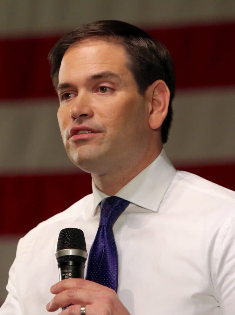 Marco Rubio speaks into a microphone in front of an American flag.