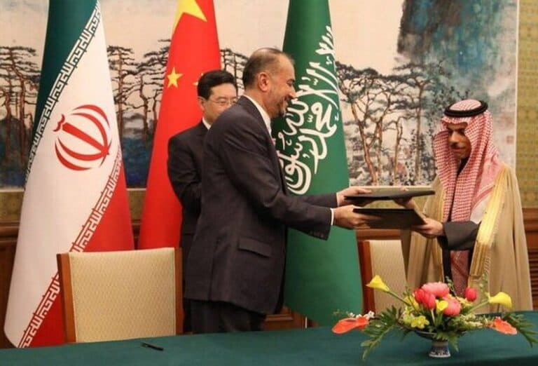 Iranian and Saudi foreign ministers exchanging documents during a diplomatic signing, with Iranian, Chinese, and Saudi flags behind them.