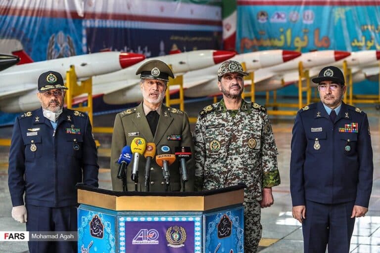 Iranian military and aviation officials stand before aircraft and missiles during a defense event in Iran.